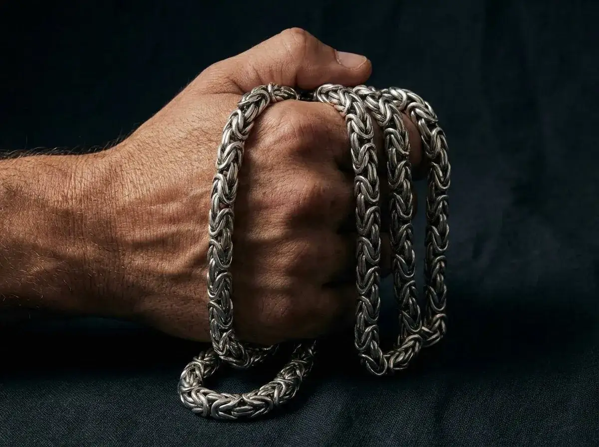 Dłoń trzyma ciężki, srebrny łańcuch o splocie przypominającym kr&oacute;lewski ankier inaczej. Biżuteria wygląda na masywną i stylową.