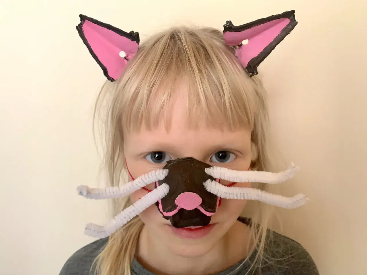 Dziewczynka z blond włosami ma na sobie uszy kota i maskę z wąsami. Idealny pomysł, jak przebrać dziecko za kota na bal przebierańców.