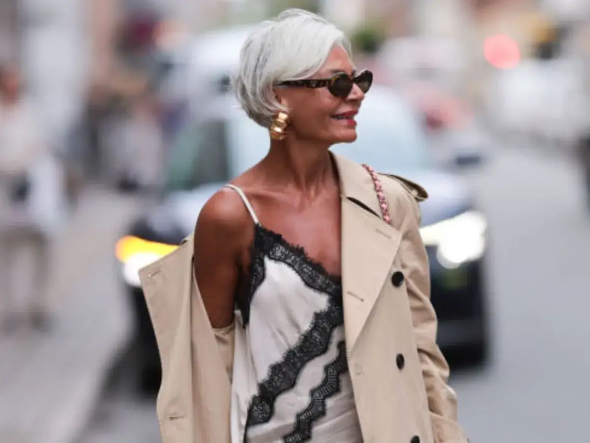 Elegancka kobieta z siwymi włosami, w beżowym płaszczu i sukience z koronką. Inspiracja, jak nosić siwe włosy z klasą.