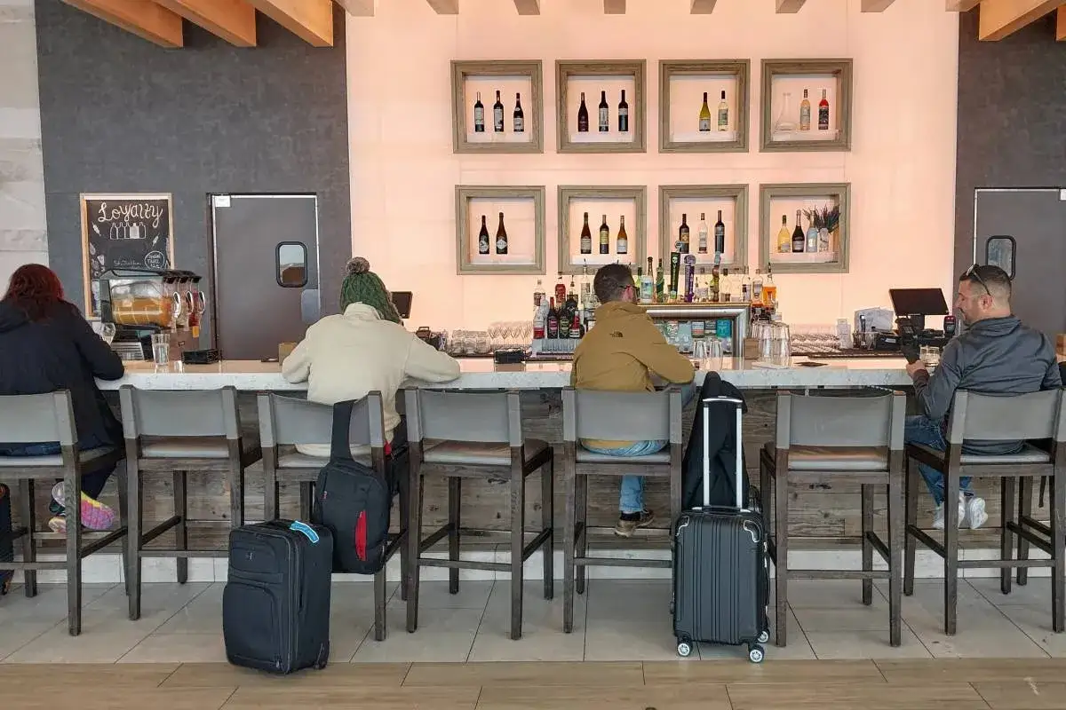 Podróżni siedzą przy barze, zastanawiając się, czy na lotnisku można pić alkohol. W tle widać butelki i sprzęt barmański.