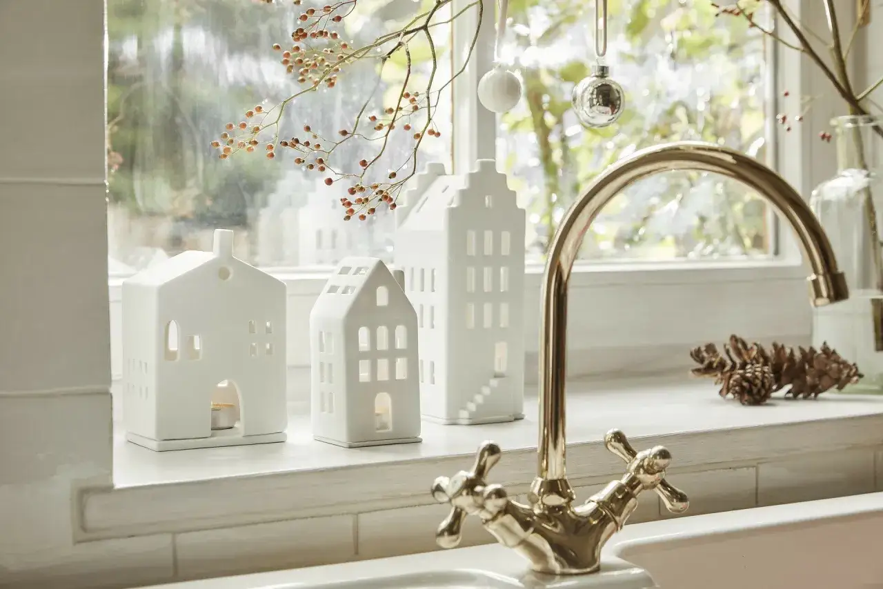 Białe ceramiczne domki tworzą urocze dekoracje okien i ozdoby na parapet. W tle gałązka z owocami i błyszczące bombki.