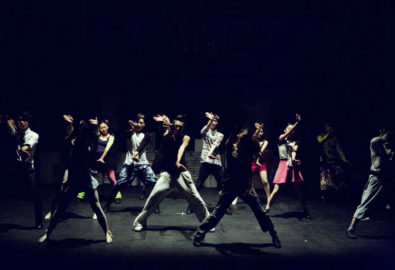 Grupa tancerzy na scenie, w ciemnościach, wykonuje choreografię. Ich ruchy tworzą dynamiczny typ tonacji.