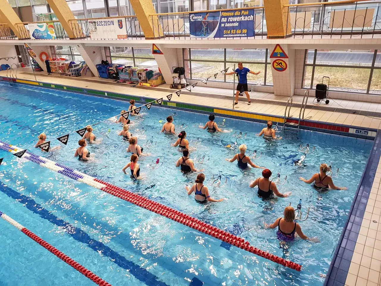 Grupa kobiet ćwiczy aqua cycling w basenie pod okiem instruktora. Woda dodaje oporu, sprawiając, że trening jest efektywny.