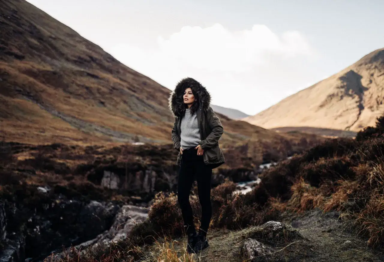 Kobieta w ciepłej, futrzanej parkce stoi na tle g&oacute;rskiego krajobrazu. To idealna odzież na chłodne dni.