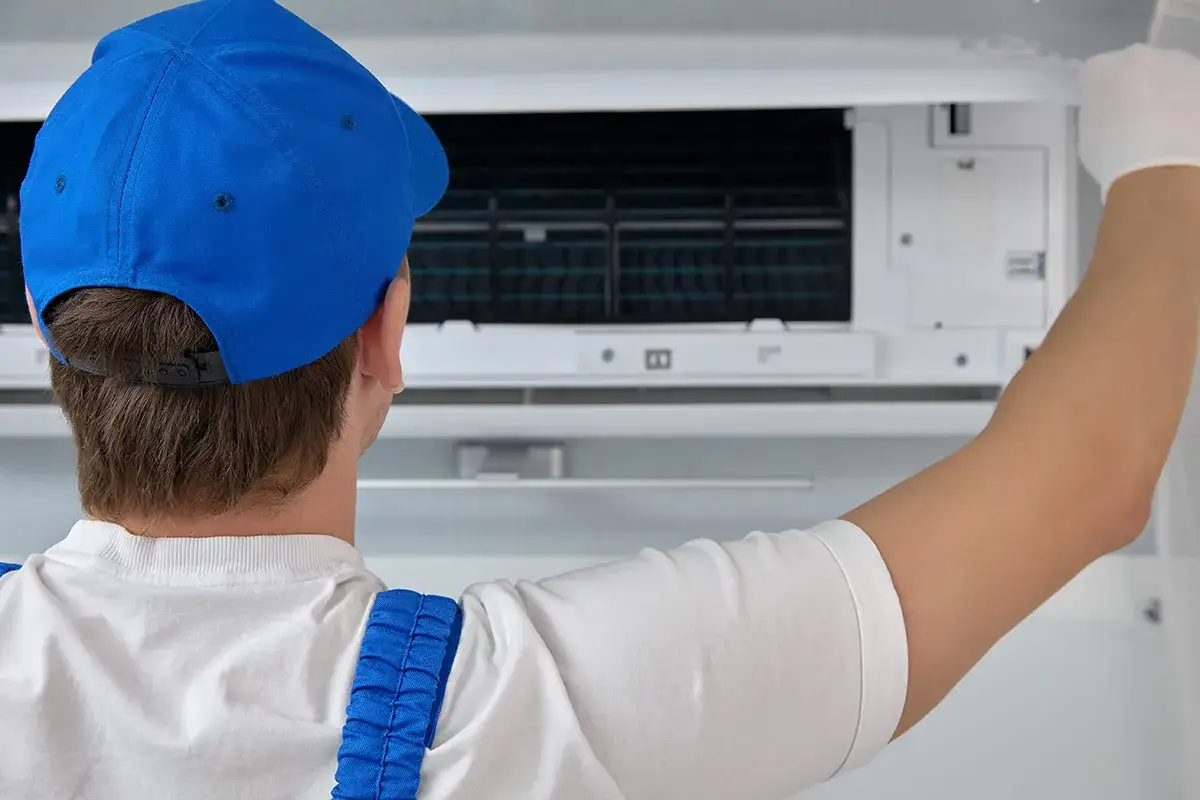 Mężczyzna w niebieskiej czapce i białej koszulce wykonuje samodzielny montaż klimatyzacji, pracując w rękawiczce.
