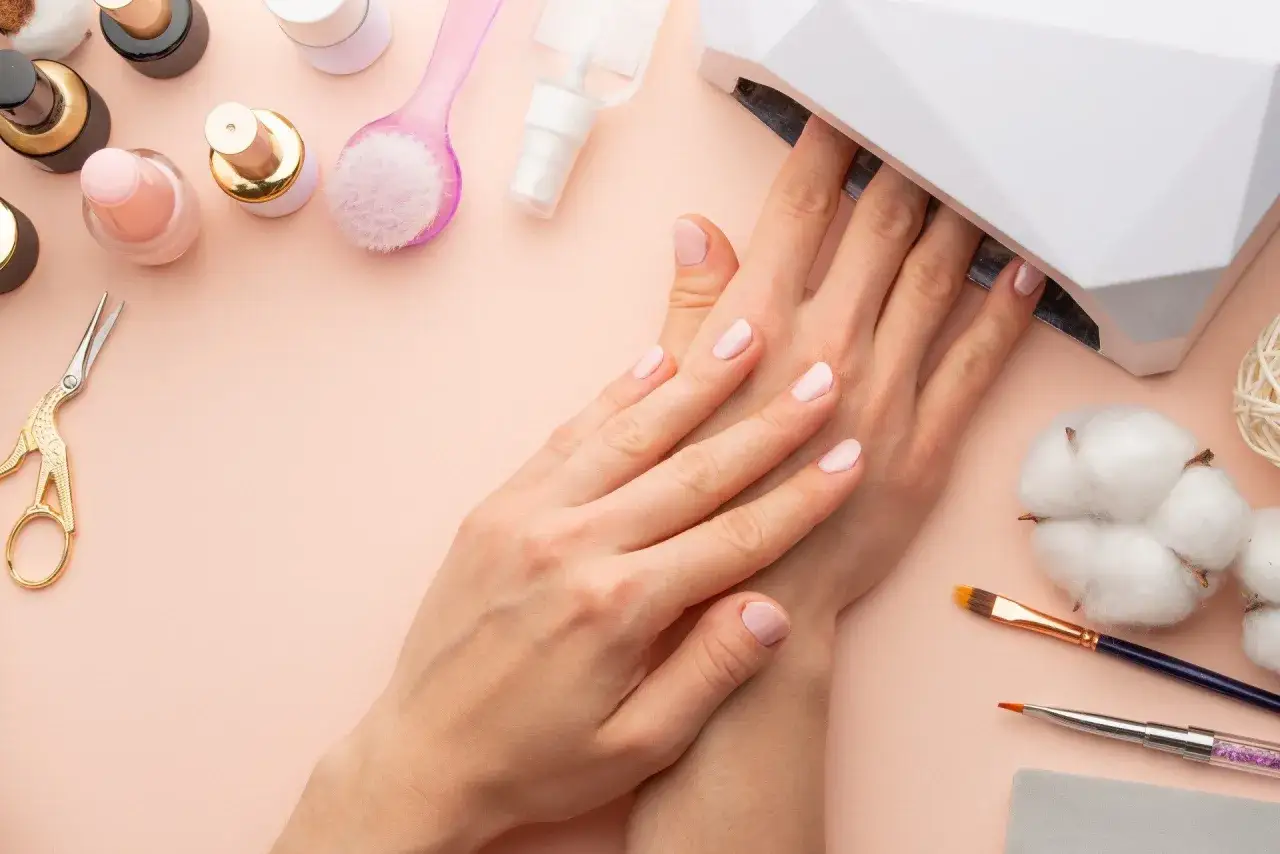 Delikatne paznokcie hybrydowe w ciąży, utwardzane w lampie UV. Wok&oacute;ł akcesoria do manicure: lakiery, pędzelki, nożyczki.