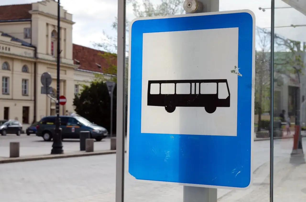 Znak przystanku autobusowego. Czy autobus wyjeżdżający z zatoki ma pierwszeństwo? W tle budynek i samochody.
