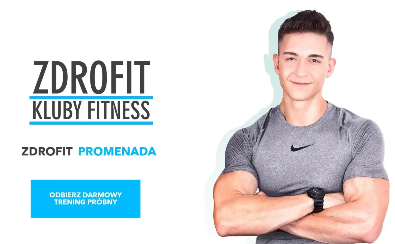 Uśmiechnięty, muskularny zdrofit trener personalny w szarej koszulce, z logo ZDROFIT KLUBY FITNESS i przyciskiem 