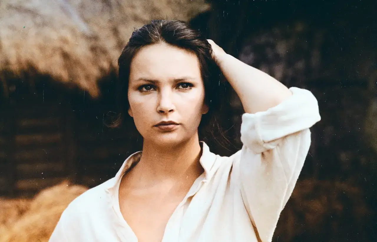 Anna Dymna w wieku 40 lat, z ręką we włosach, w prostej, lnianej koszuli, na tle wiejskiego krajobrazu.