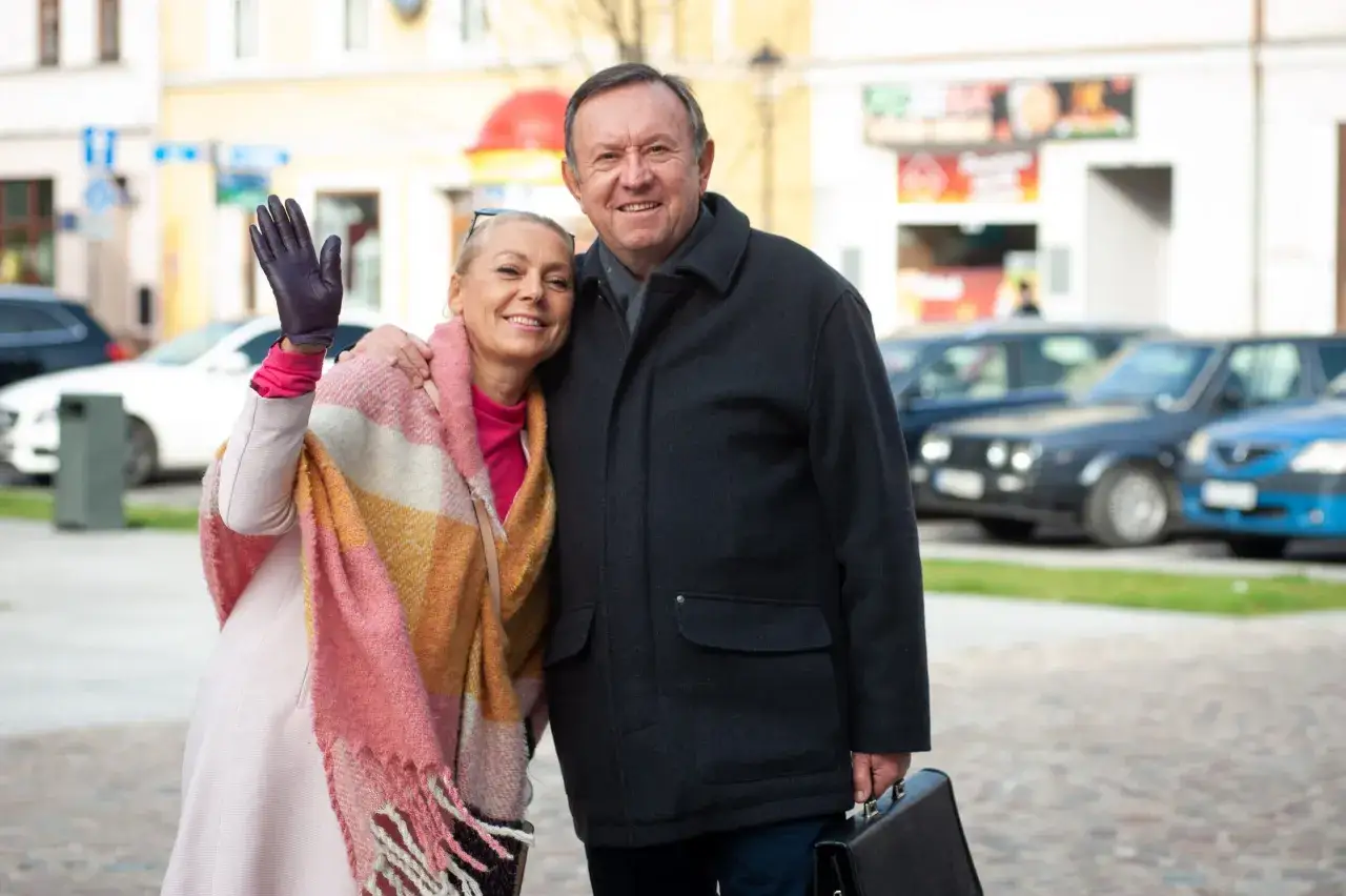 Uśmiechnięta Małgorzata Szeptycka w kolorowym szalu pozdrawia, obejmując partnera z teczką.