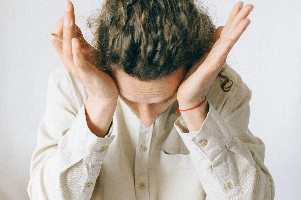 Mężczyzna z kręconymi włosami trzyma dłonie przy skroniach, pochylając głowę. Czuje b&oacute;l oka przy poruszaniu gałką.