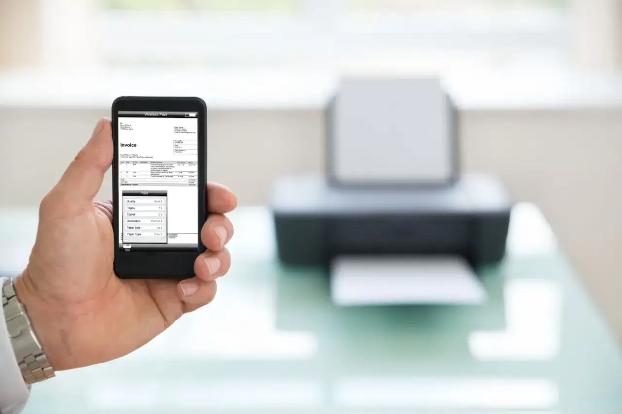 Dłoń trzyma smartfon z fakturą, obok drukarka. Prosty spos&oacute;b na to, jak drukować z telefonu.