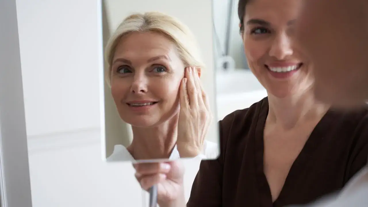 Uśmiechnięta 40-latka w lustrze, dotykająca swojej twarzy. Pokazuje, jak powinna wyglądać twarz 40-latki, pełna naturalnego blasku i pewności siebie.