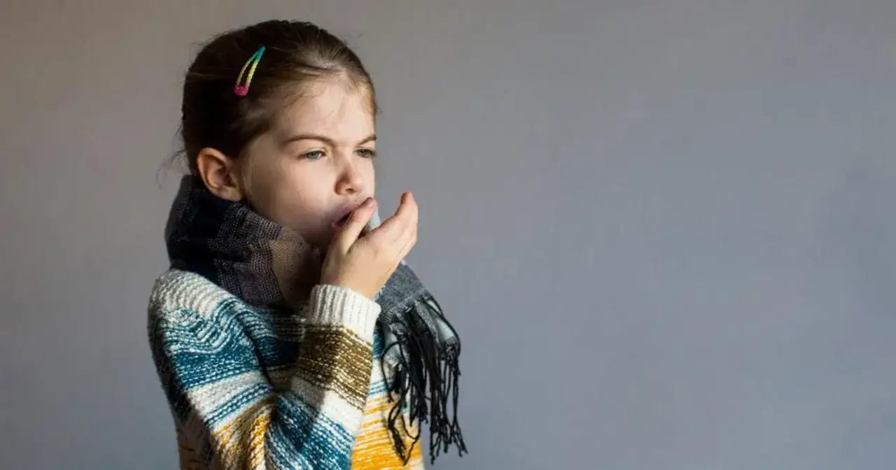 Dziewczynka w szaliku i swetrze zakrywa usta dłonią, kaszel mokry męczy ją od rana.