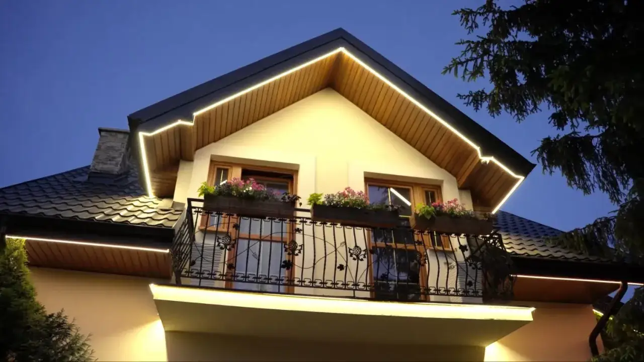 Elegancki dom nocą, z ciepłym światłem led w podbitce podkreślającym architekturę. Balkon z kwiatami i ozdobną balustradą.