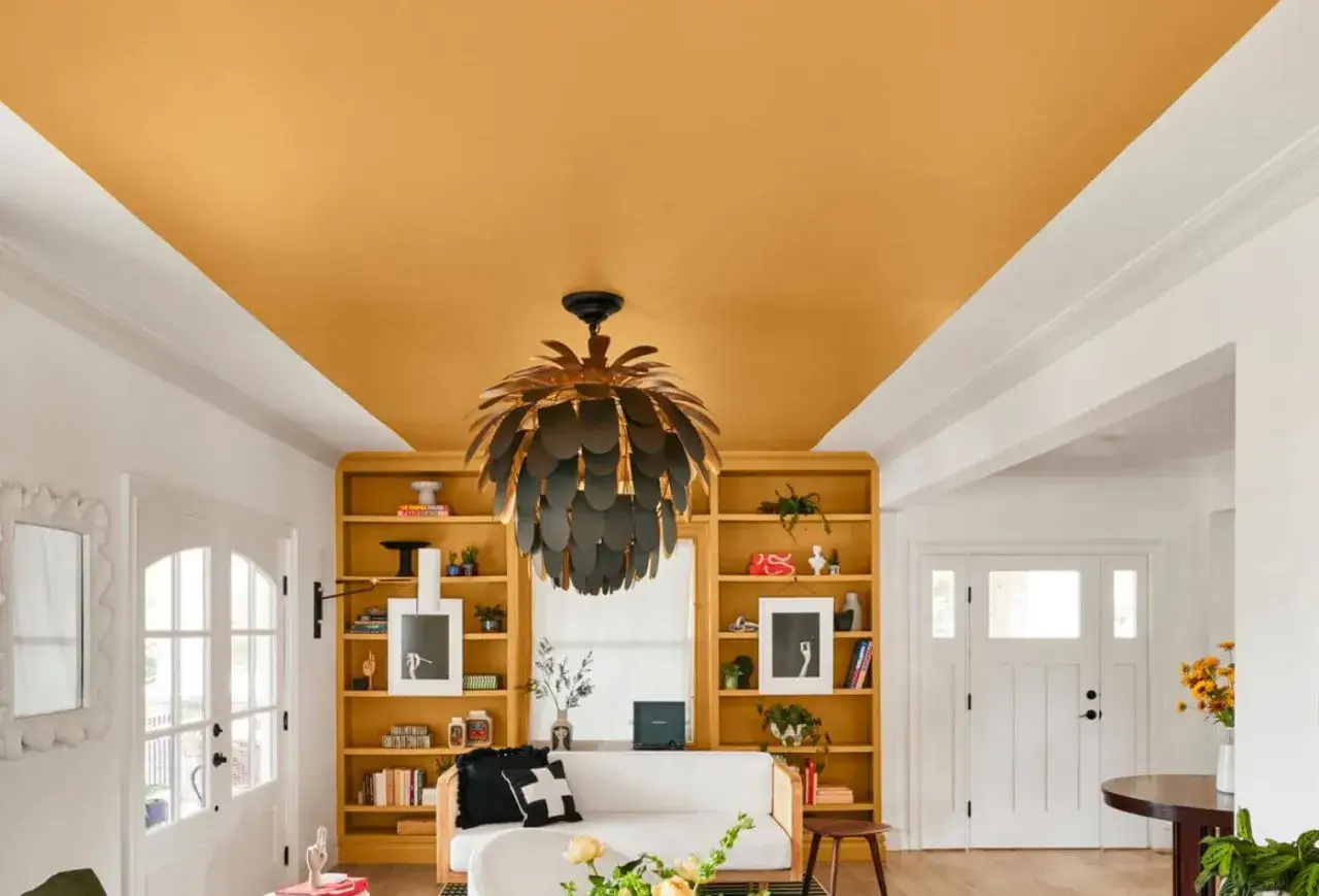 Jasny sufit i ż&oacute;łte regały to trik, jak optycznie powiększyć pok&oacute;j kolorami. Wnętrze z designerską lampą i białymi drzwiami.