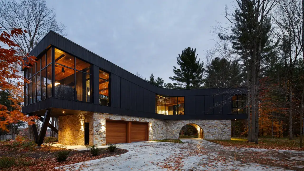 Nowoczesny dom z kamienia i drewna, z czarną, falistą bryłą na kamiennym cokole, otoczony jesiennym lasem.