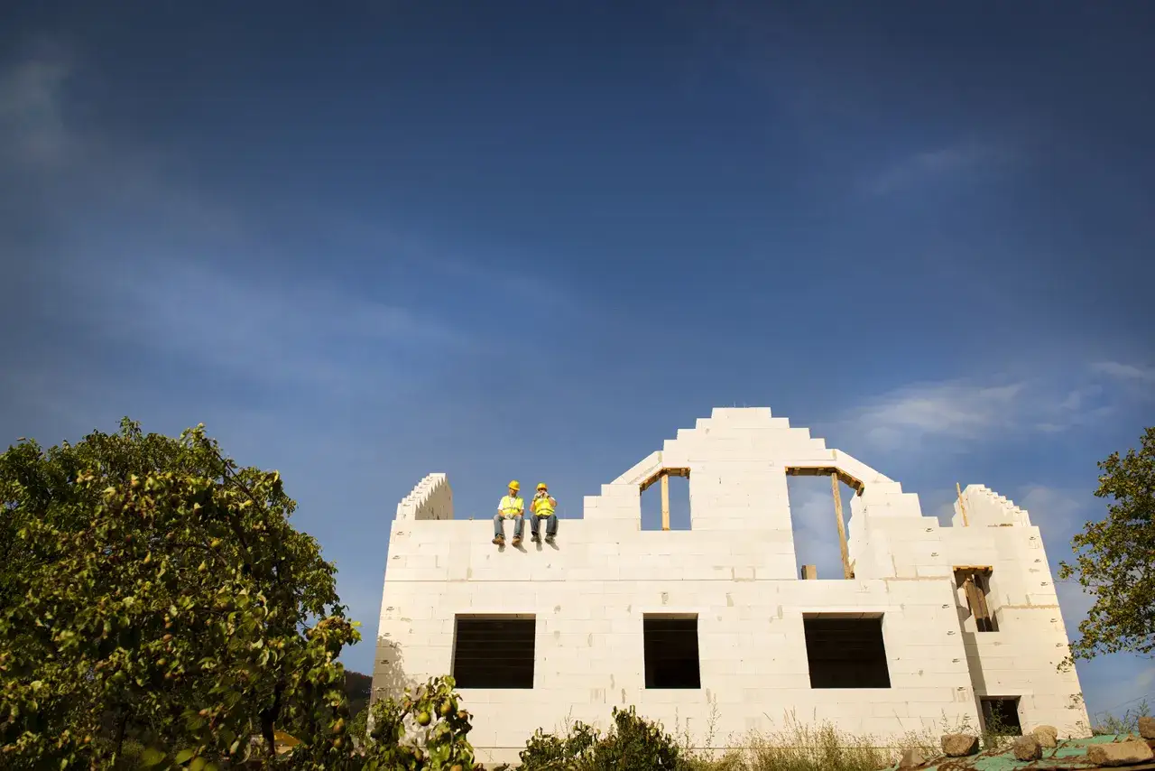 Dwóch budowlańców w kaskach siedzi na dachu domu w trakcie budowy. W tle błękitne niebo.