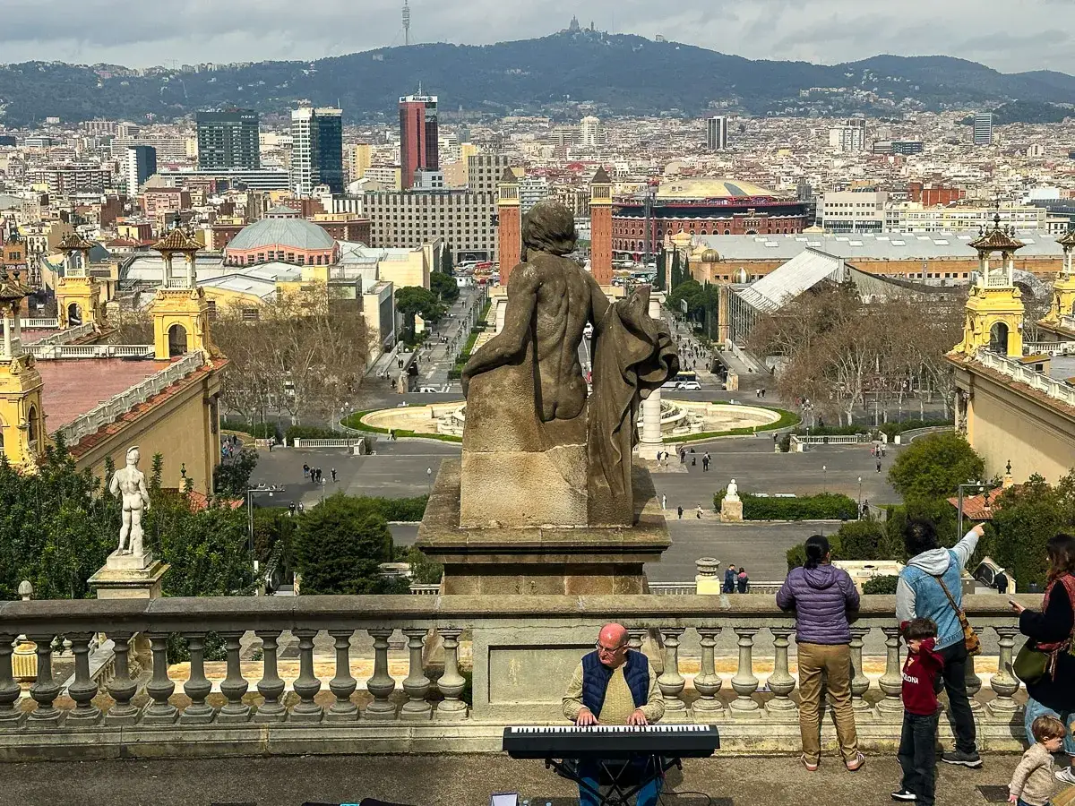 Widok na Barcelonę z tarasu Muzeum Narodowego Katalonii. Na pierwszym planie grający muzyk i rodzina, w tle panorama miasta.