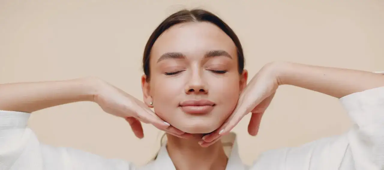 Kobieta z zamkniętymi oczami wykonuje ćwiczenia jogi twarzy, delikatnie masując podbr&oacute;dek i policzki dłońmi.