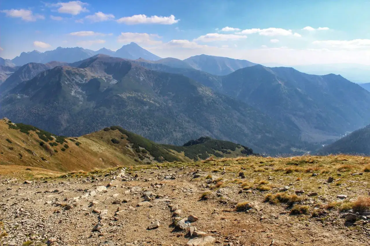 Kamienista ścieżka prowadzi przez trawiaste zbocze ku pasmu g&oacute;rskiemu. W oddali widać majestatyczne Czerwone Wierchy, kt&oacute;rych wysokość zapiera dech w piersiach.