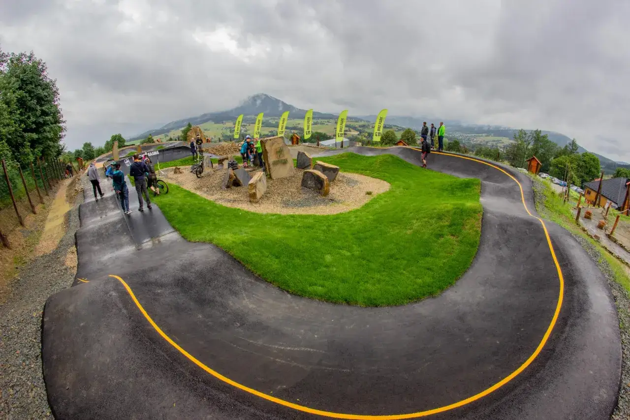 Ludzie na rowerach w bike parku Kasina, z pagórkami i zieloną trawą. W tle góry i chmury.