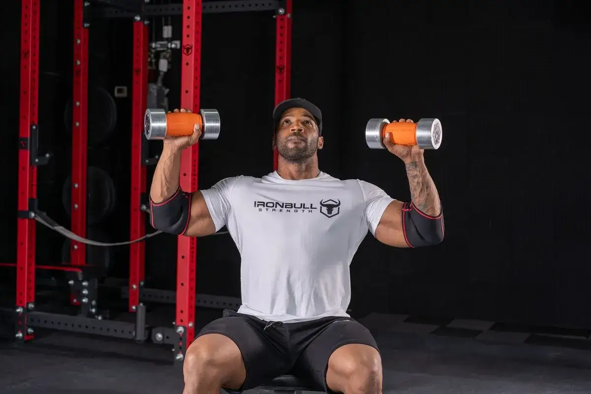 Mężczyzna w czapce i koszulce Ironbull Strength wykonuje trening chwytu z hantlami.