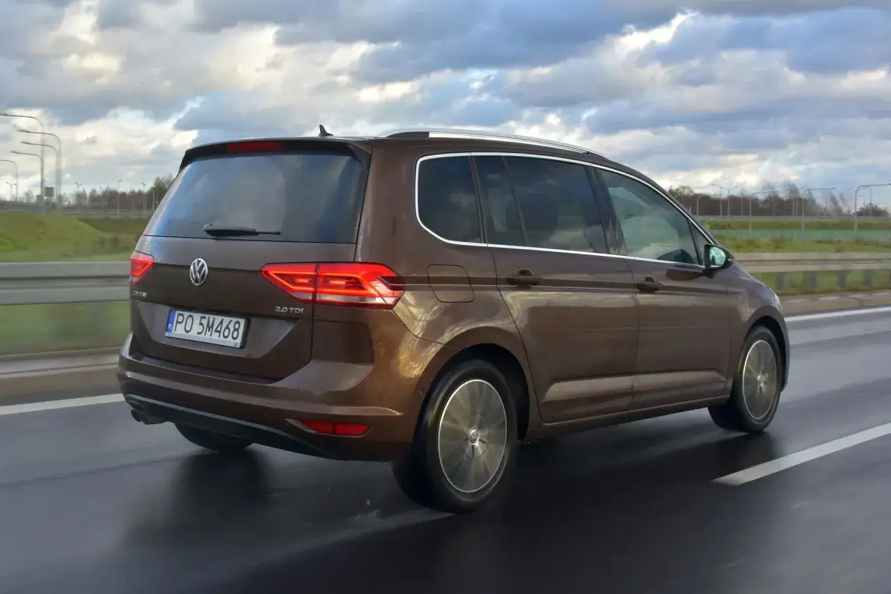 Brązowy Volkswagen Touran 2.0 TDI jedzie po mokrej drodze pod pochmurnym niebem.