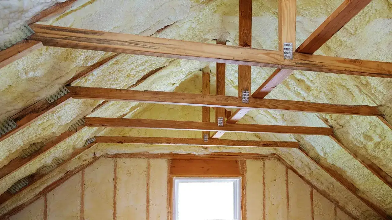 Drewniana konstrukcja poddasza pokryta żółtą pianką do ocieplenia. Widać okno i belki stropowe.