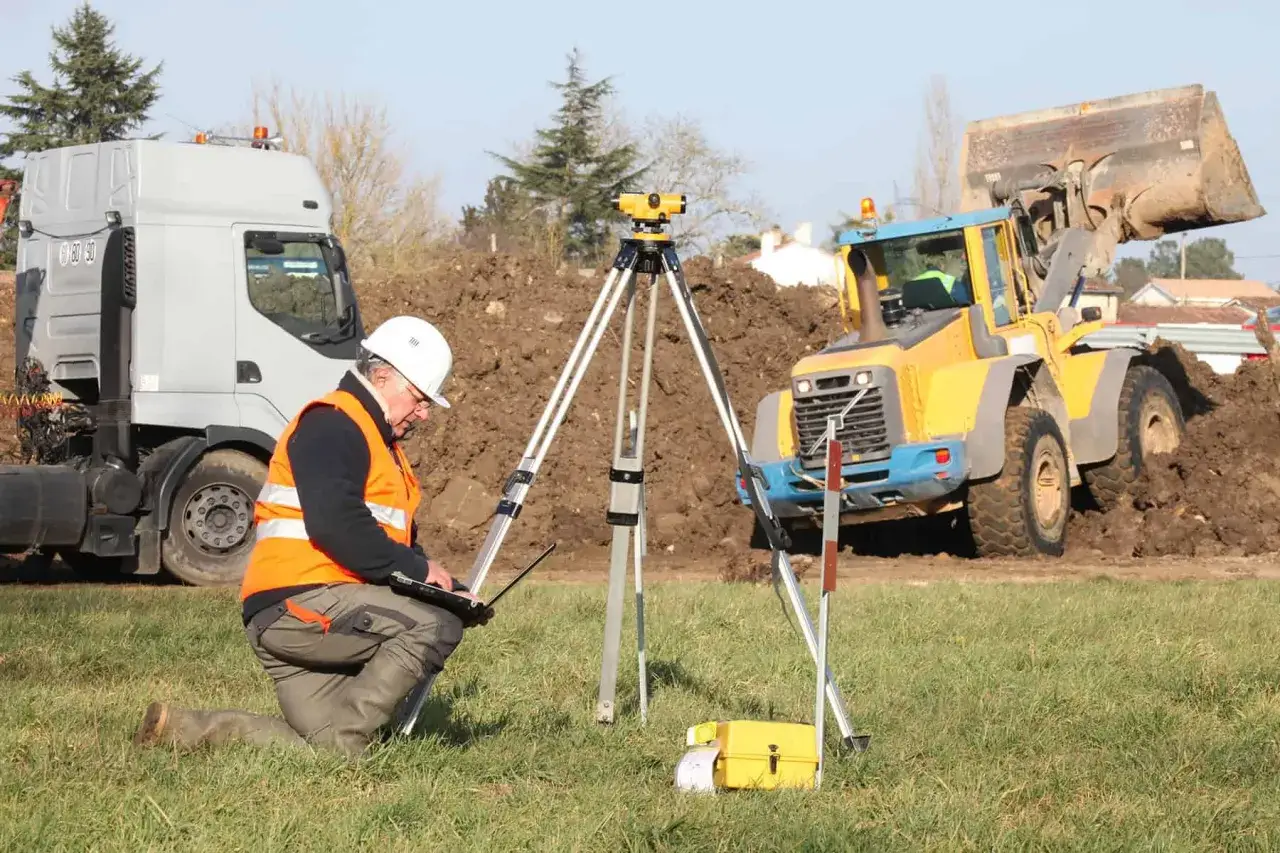 Geodeta wykonuje badanie gruntu pod budowę domu, korzystając z niwelatora i laptopa. W tle koparka i ciężarówka.