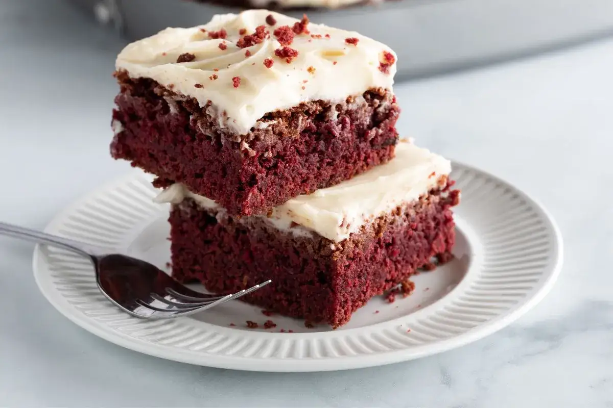 Dwa kawałki ciasta red velvet z kremowym lukrem i posypką na talerzyku z widelcem.