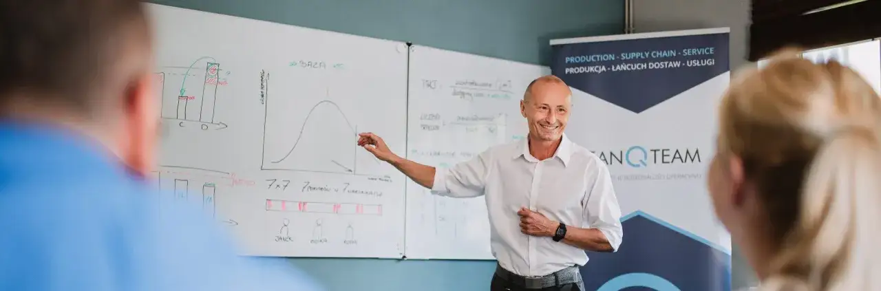 Mężczyzna w białej koszuli tłumaczy wykresy na tablicy. W tle baner z napisem 