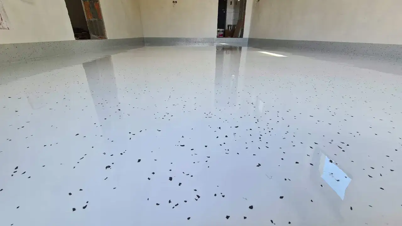 Lśniąca, biała posadzka żywiczna do garażu z czarnymi płatkami. Wygląda jak nowa, gotowa na przyjęcie samochodu.