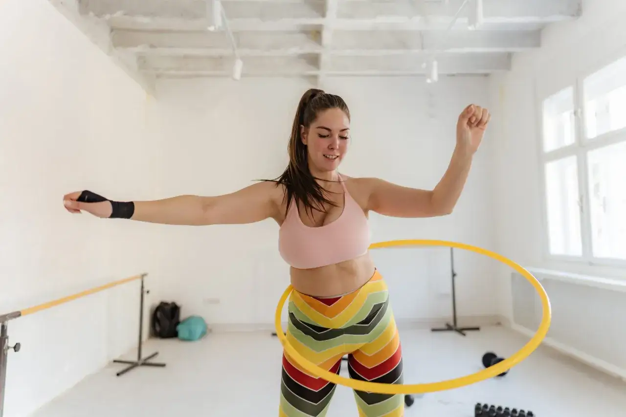 Kobieta ćwiczy z hula hop, zastanawiając się, czy hula hop odchudza. Wesoła atmosfera treningu w jasnej sali.