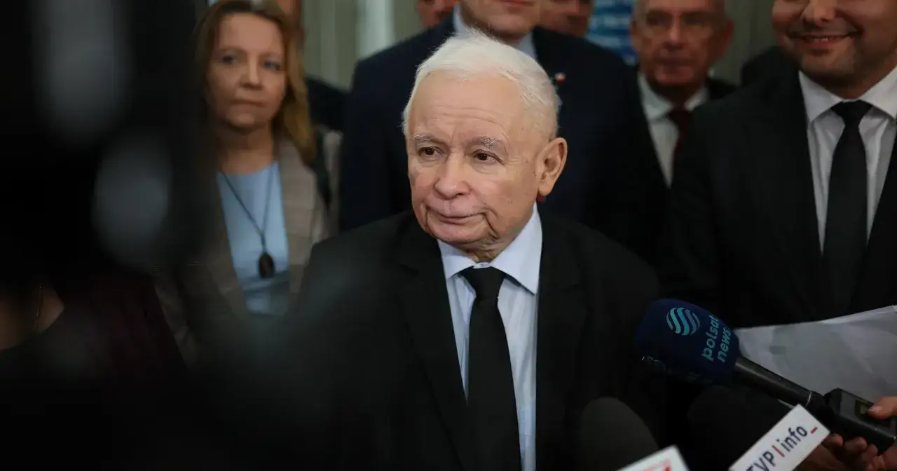Jarosław Kaczyński w otoczeniu innych polityków, udziela wywiadu. W tle widać kobiety i mężczyzn, a przed nim mikrofony mediów.