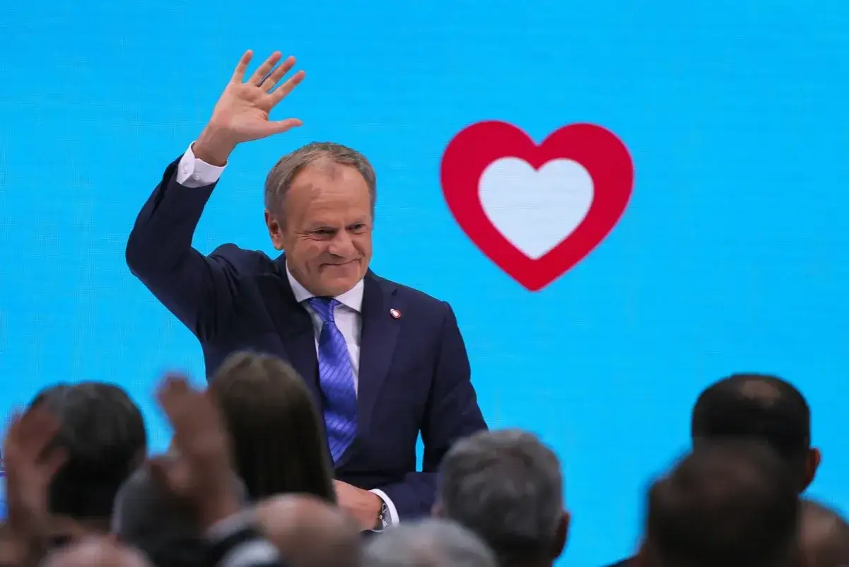 Donald Tusk pozdrawia tłum na tle niebieskiego ekranu z sercem i logo Koalicji Obywatelskiej.
