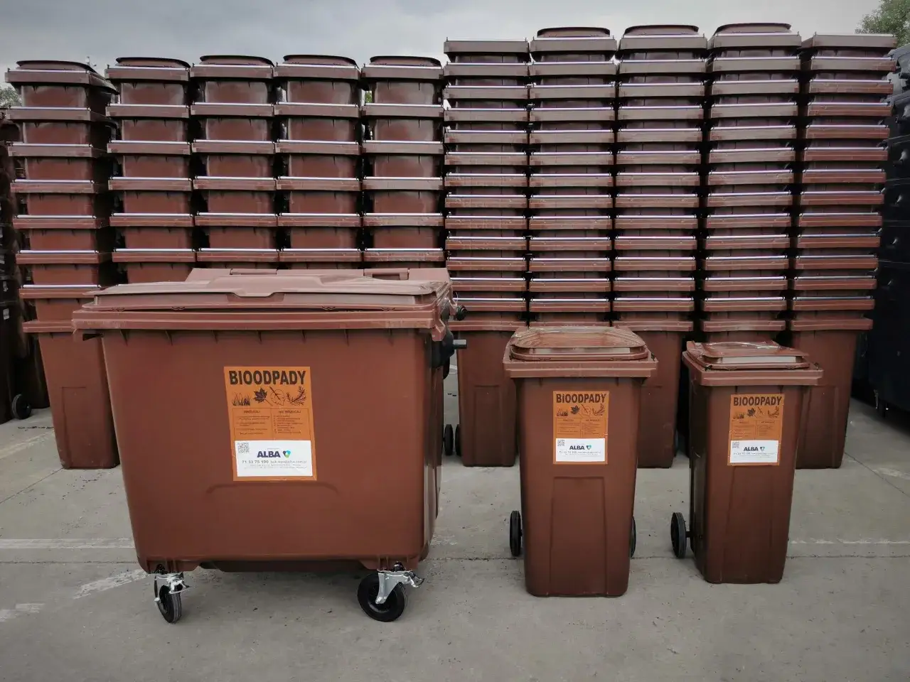 Brązowe pojemniki na bioodpady, jeden duży i dwa mniejsze, stoją przed stosem identycznych, zamkniętych kontenerów. Na jednym z nich widnieje napis 