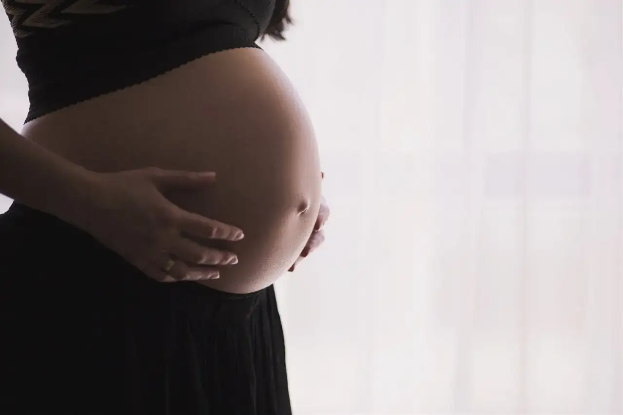 Dłońmi obejmuje zaokrąglony, twardy brzuch w ciąży. Delikatne światło podkreśla jego kształt.