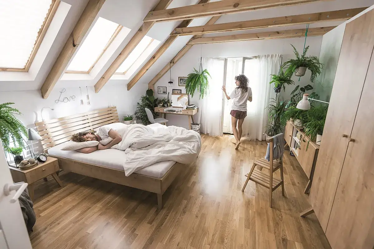 Sypialnia na poddaszu z ł&oacute;żkiem, na kt&oacute;rym śpi mężczyzna. Obok ł&oacute;żka stolik nocny. W tle kobieta przy oknie. W pokoju dużo roślin. Ścianka kolankowa 140 cm.