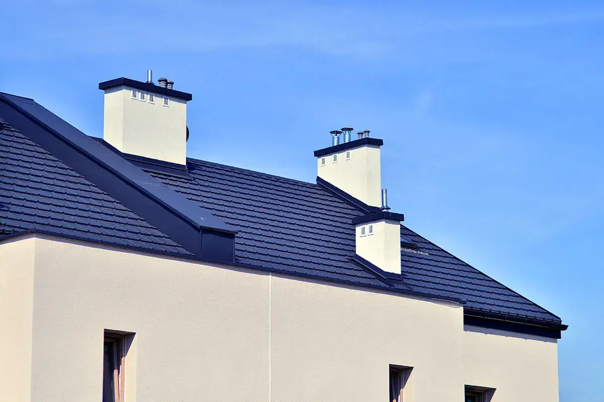 Nowoczesny dach bez okapu z ciemnogranatową dachówką i białymi kominami na tle błękitnego nieba.