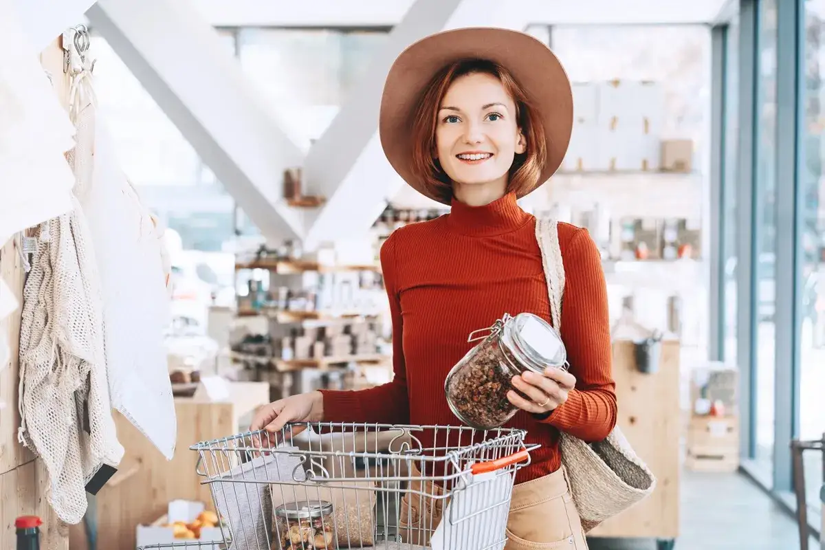 Uśmiechnięta kobieta z koszykiem i słoikiem pełnym granoli. Unika skutków kupowania produktów z dołu rankingu opłacalność vs jakość.