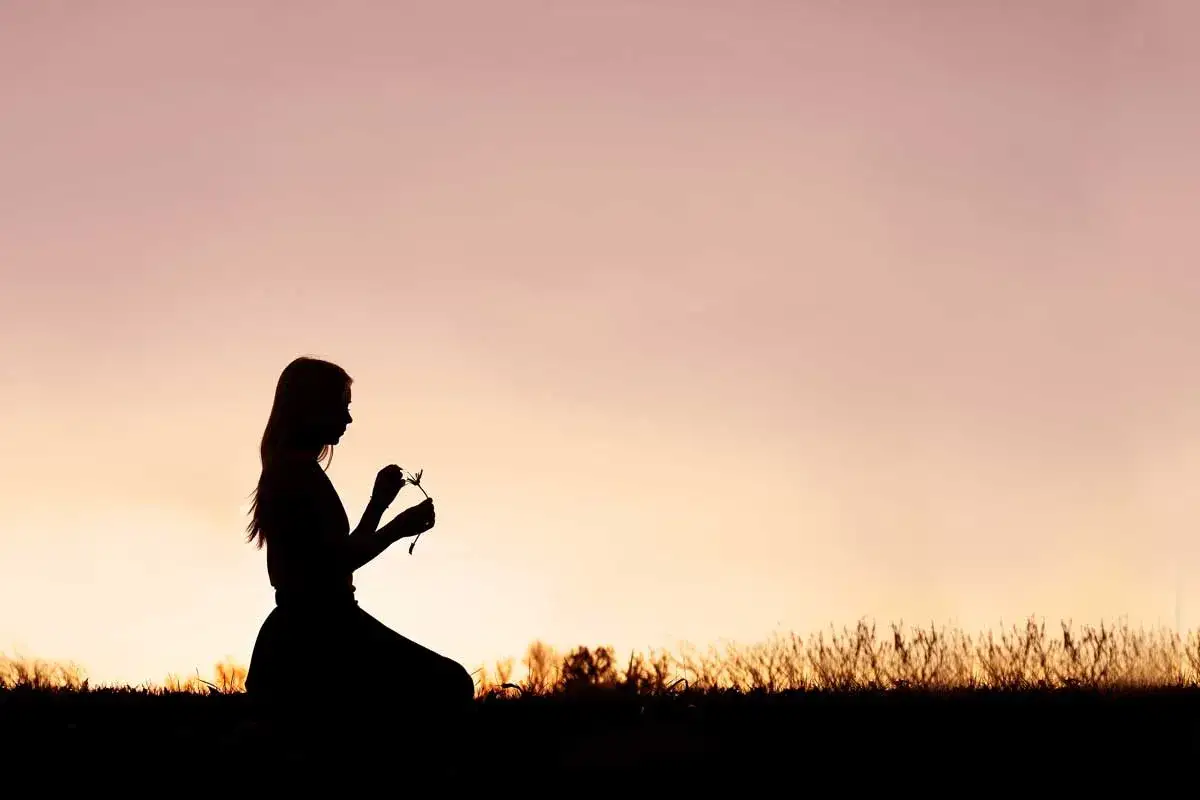 Sylwetka dziewczynki klęczącej na trawie, trzymającej w dłoniach mały kwiatek. W jej postawie widać tęsknotę za kimś, z kim nie można być.