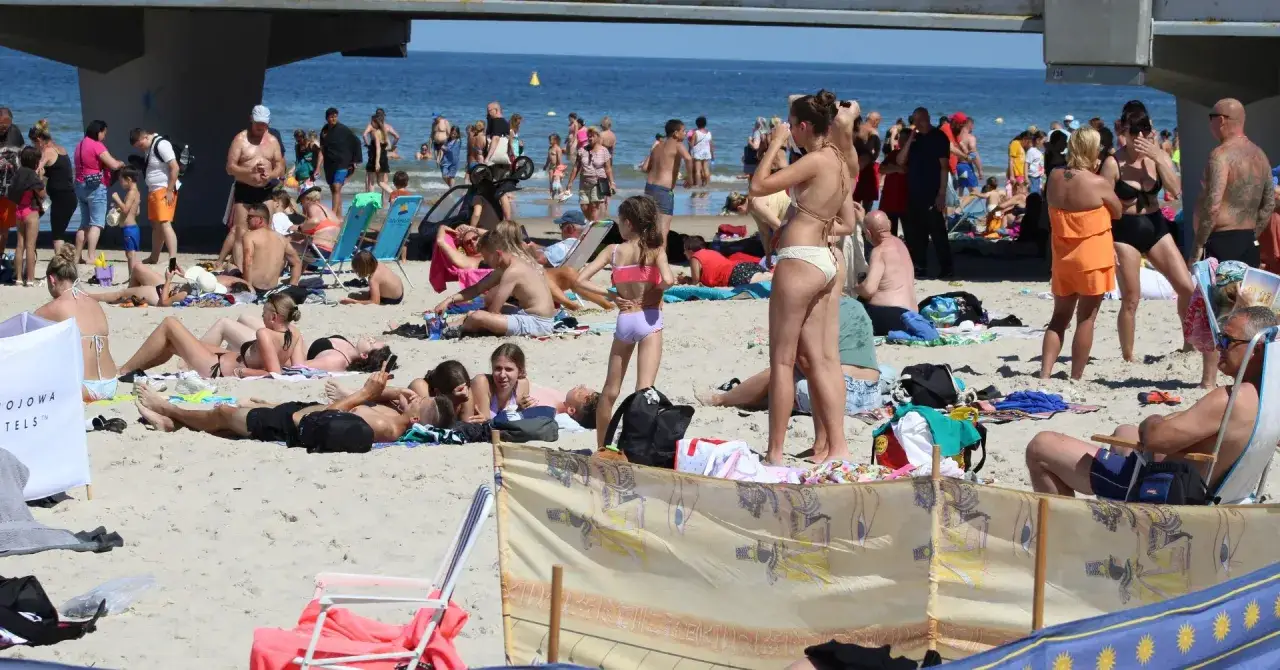 Słoneczny dzień na kołobrzeskiej plaży. Ludzie odpoczywają na piasku, kąpią się w morzu i spacerują pod molo.