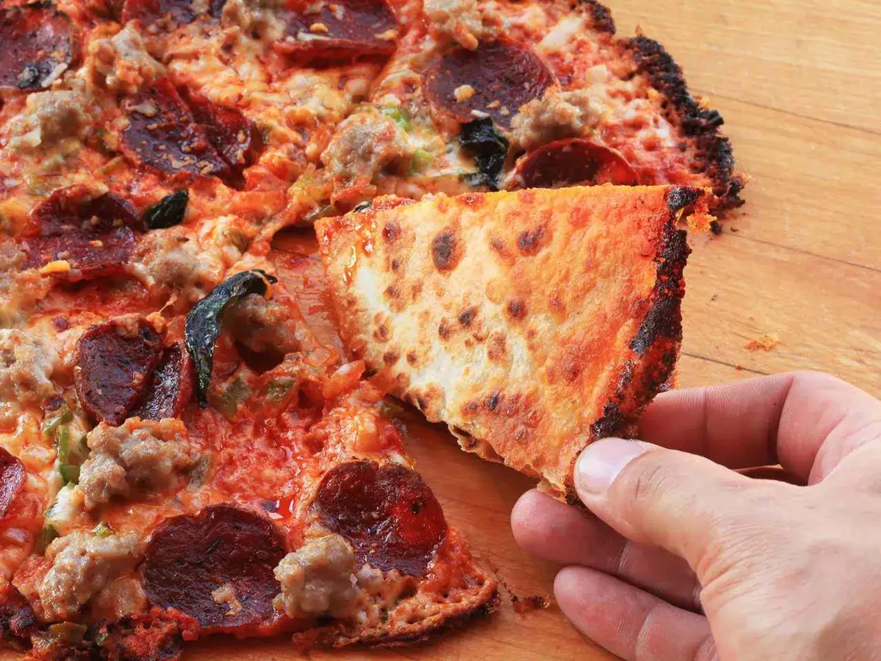 Chrupiąca pizza z tortilli z serem, pepperoni i kiełbasą, ręka podnosi kawałek.