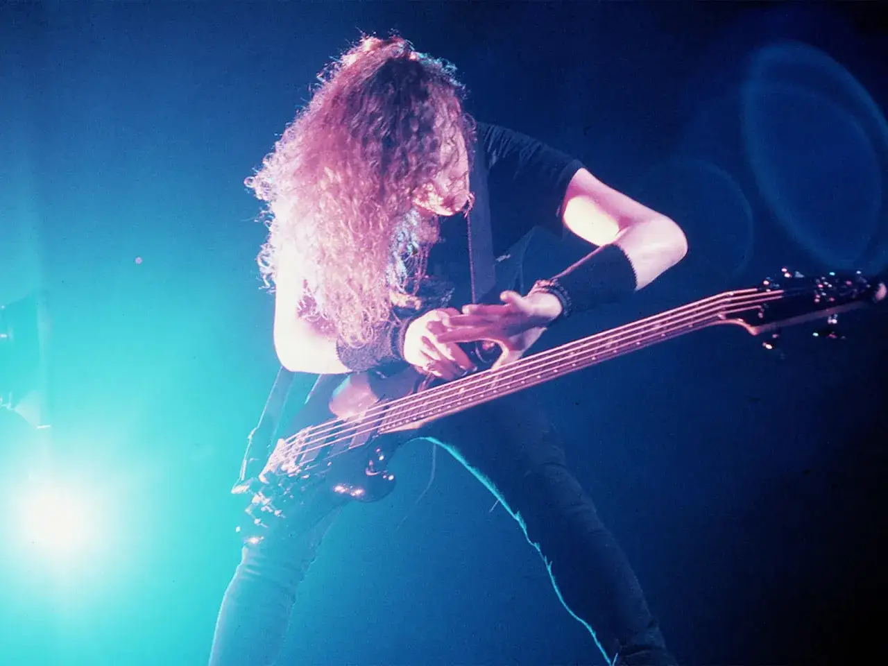 Jason Newsted z zespołu gra na basie pod niebieskim światłem. Jego długie, kręcone włosy opadają na twarz.