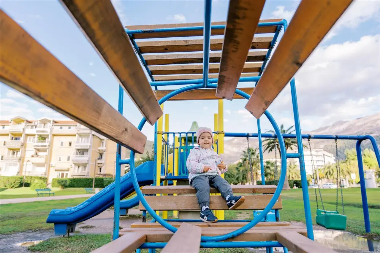Mała dziewczynka siedzi na ławce na placu zabaw, trzymając pluszową zabawkę. Od kiedy otwarte place zabaw dla dzieci, takie widoki są radością.