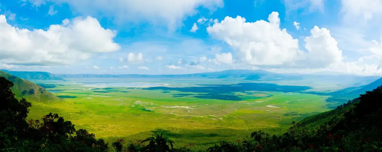 Rozległa panorama Ngorongoro Crater z zielonymi równinami, jeziorem i błękitnym niebem z chmurami.