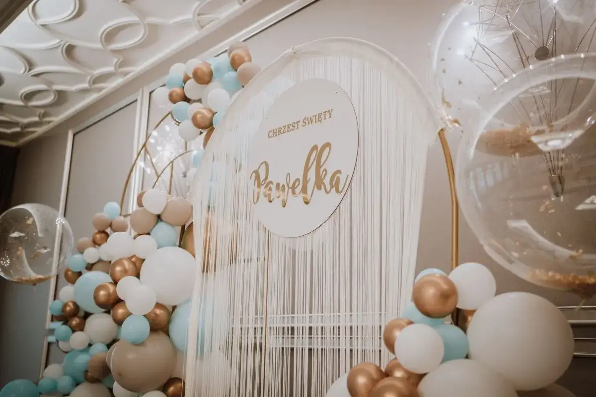 Dekoracja na chrzciny z imieniem Pawelka. Balony w kolorach bieli, złota i błękitu tworzą tło dla uroczystości.