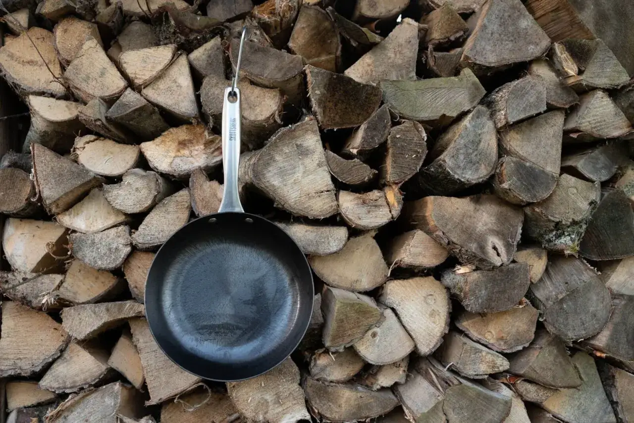 Patelnia żeliwna wisi na tle stosu drewna. Dowiedz się, jak przygotować patelnię żeliwną do pierwszego użycia.
