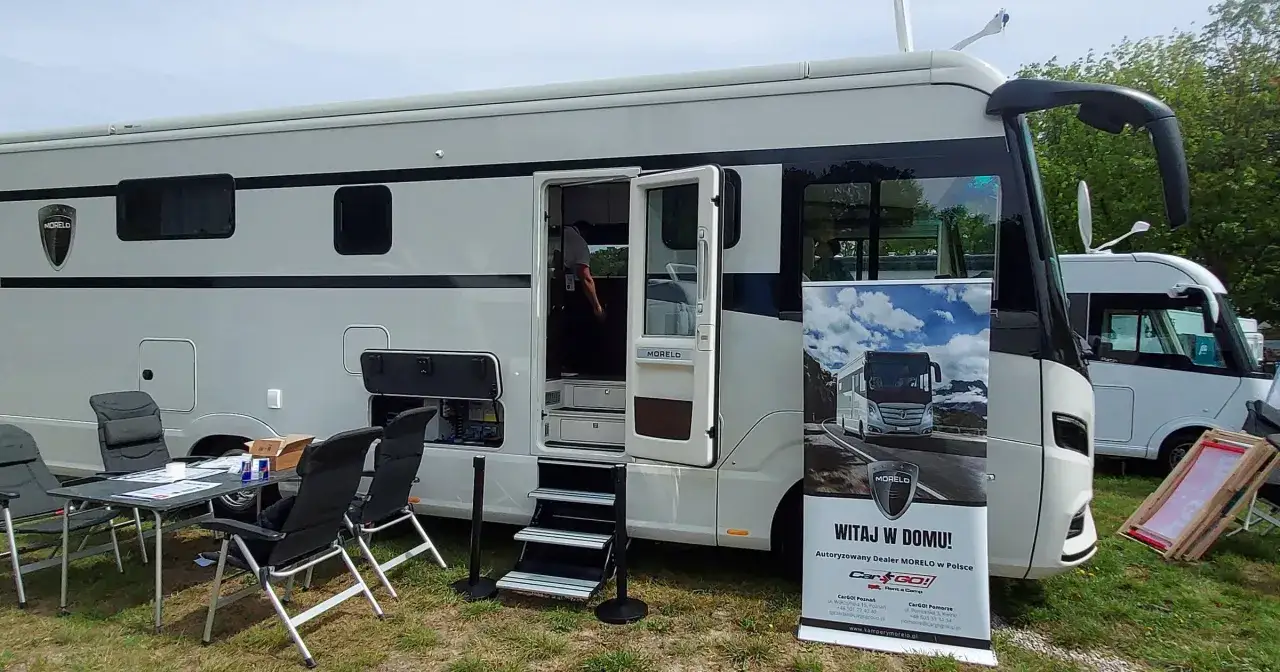 Duży kamper kat C marki Morelo z otwartymi drzwiami i schodkami. Obok stolik z krzesłami i baner reklamowy.